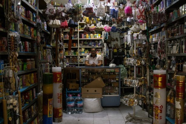 Comerciante atrás do balcão de uma pequena loja repleta de produtos.